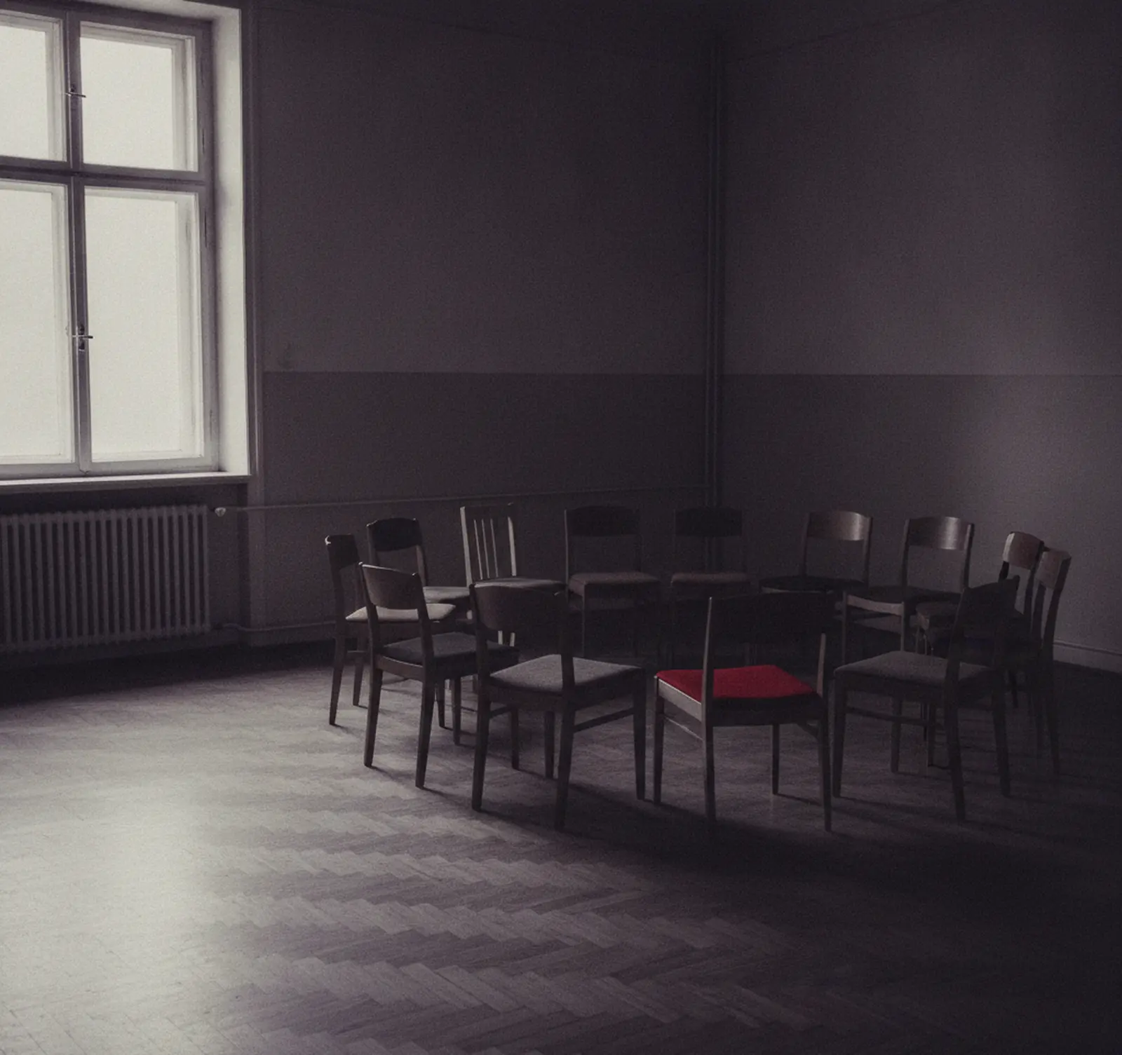 Ein leerer Stuhlkreis in einem schlichten Gemeinschaftsraum mit hohem Altbaufenster, ein Stuhl rot gepolstert